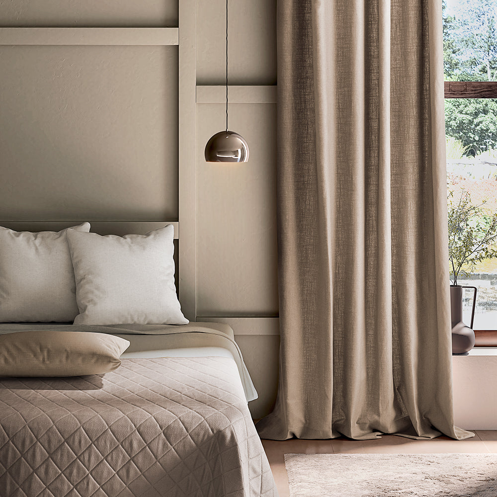 Neatly made bed with beige bedding and pillows in a bedroom with curtains and a lamp.