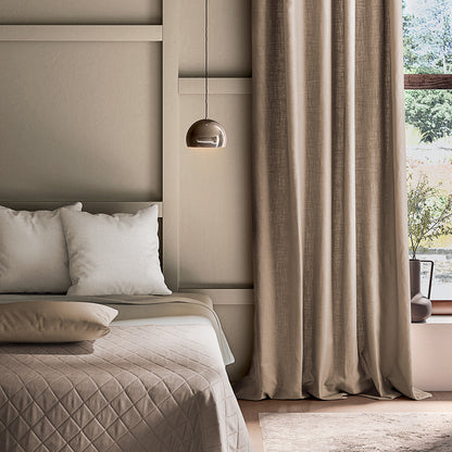 Neatly made bed with beige bedding and pillows in a bedroom with curtains and a lamp.
