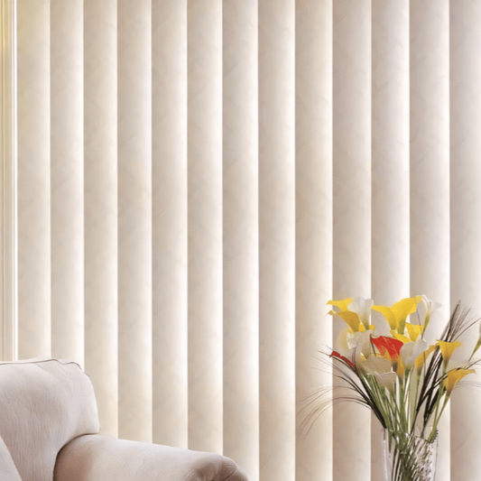 A set of white vertical blinds covering a window with a vase of colorful flowers and a white chair in the foreground.